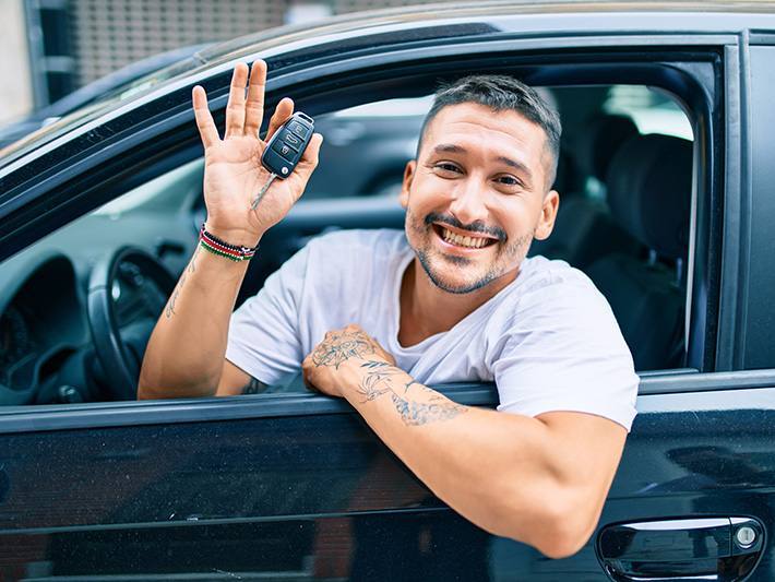 Aus dem geöffneten Fahrerfenster eines Autos beugt sich mit dem Autoschlüssel in der Hand ein strahlender junger Mann