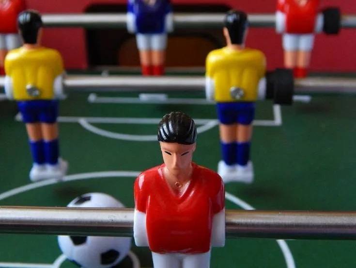 Bildausschnitt eines Tischkicker-Spielfeldes mit Spielfiguren in gelben und roten Trikots an Stangen und dem Ball im Spielfeld